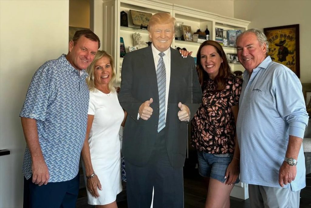 Group of four adults posing indoors beside a life-size cardboard cutout of a suited political figure.